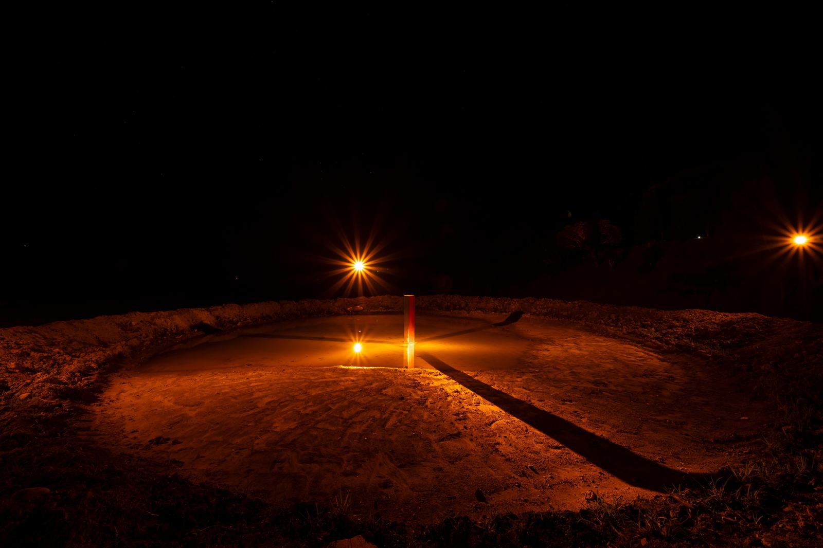 夜の円形の敷地の中心に立つ標と橙の灯り。インスタレーション作品の遠景